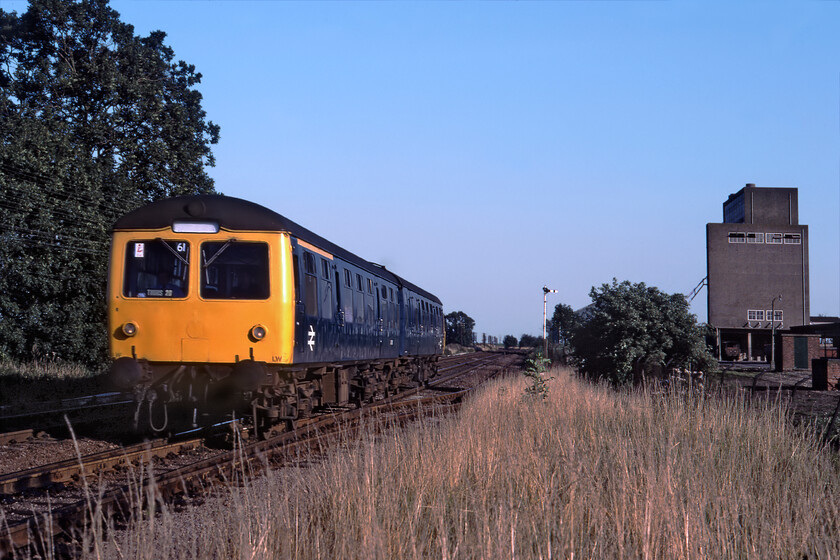 Class 105, 18.07 Ely-Stamford, Chetisham 
 A two-car Cravens Class 105 DMU catches some soft evening light as it approaches the level crossing at Chettisham. It is working the 18.05 Ely to Peterborough service, a relatively short service across the Fens via March. The large building to the right in this view is a rail-connected grain storage depot that saw pretty regular traffic. Before this use, it was a Government storage depot. There is a local story that when its use as a grain store came to an end and the whole site was demolished boxes of margarine dating from World War 2 were found in deep storage on site. 
 Keywords: Class 105 DMU 18.07 Ely-Stamford Chetisham First generation dmu