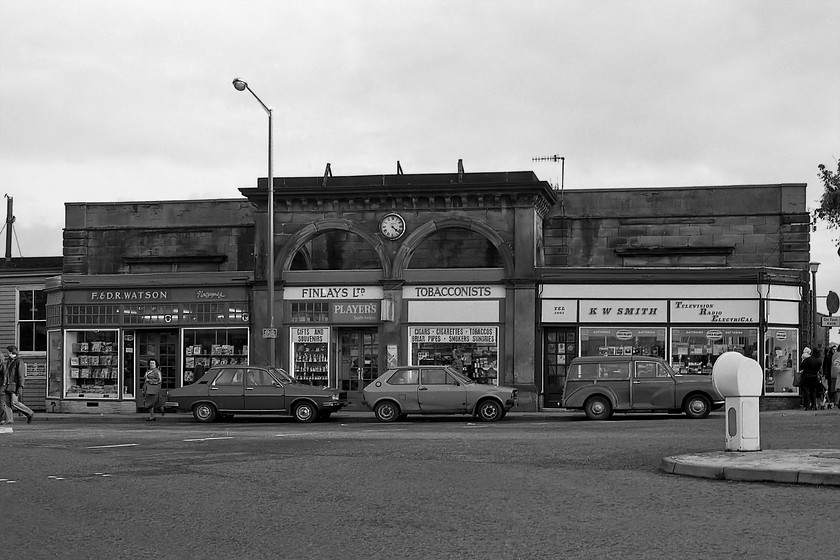 Station frontage (LNER clock-working!), Whitby station 
 Behind these rather crudely grafted on shop fronts is a railway station! The giveaway is the LNER clock above the former station entrance that was working and reading the correct time. Whitby station was designed by George Townsend Andrews for the York and North Midland Railway (Y&NM) and was opened in 1845. Today Messrs Watson, Finlays and Smith and their premises have all moved on revealing the original frontage of this fine Grade II listed station. The entrance has reverted to the front again through the twin arches rather than the rather lowly entrance being used at this time. Three period cars are seen parked in the twenty minutes bays, a facelift Renault 12, a rather beaten up VW Polo that would only be up to four years old and a classic Morris Minor 'woody'. I visited the station in 2019 with no cars parked in front now and by way of comparison, I took a similar photograph, see..... https://www.ontheupfast.com/p/21936chg/26273779804/frontage-whitby-station Interestingly, during this visit the clock was not working! 
 Keywords: Station frontage LNER clock Whitby station