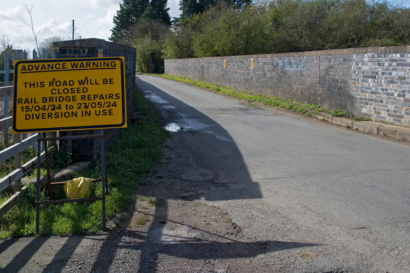 Closure sign, Bridge 204, Ashton Road 
 Bridge 204 crosses the WCML between the Northamptonshire villages of Roade and Ashton. The bridge was constructed in the 1960s when the line was electrified replacing the original structure that could not offer the required clearance for the wiring. Unfortunately, like many structures dating from this era that adopted the new building techniques of the time they have not fared well and this bridge is no exception. Being what is, essentially, a slab of concrete with brick-built parapets placed on top it has shifted and the parapets are now teetering requiring reconstruction. The eastern end was worked on a couple of years ago with the repaired portion seen to the extreme right in the image but the parapet at the western end is now requiring urgent attention necessitating another lengthy road closure as seen here from the road sign. 
 Keywords: Closure sign Bridge 204 Ashton Road