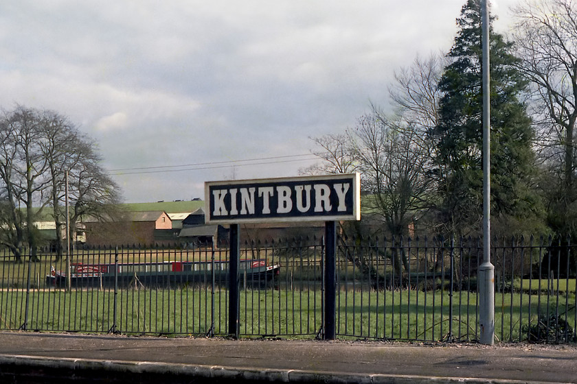 Running-in board, Kintbury station 
 A lovely GW running-in board at Kintbury station mounted on posts made of old track. In the background a pretty narrow boat sits moored up on the Kennet and Avon canal. 
 Keywords: Running-in board Kintbury station