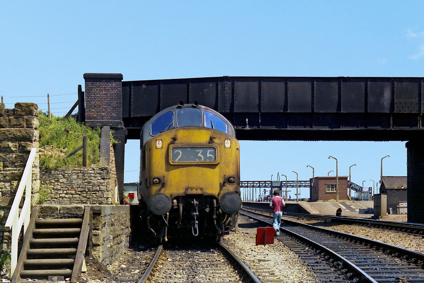 37235, stabled, Severn Tunnel Junction MPD 
 37235 stands under the bridge with Severn Tunnel Junction station in the background. It is displaying its number in the now defunct head code panel. This often proved useful for us spotters when the usual white cab side number was tricky to read. 37235 had moved west to be based at Landore. It resided here for sometime remaining a West Country engine for some years before moving back up north again. It had an ignominious end being put in store as 'unserviceable' in 1995. It then went through several owners, was subject to component recovery (stripping to you and I) and finally cut up by WCRC at Carnforth in 2008. Notice one of my friends calmly walking along the side of a running line. I remember that I did not take this route but went up the steps to the left!