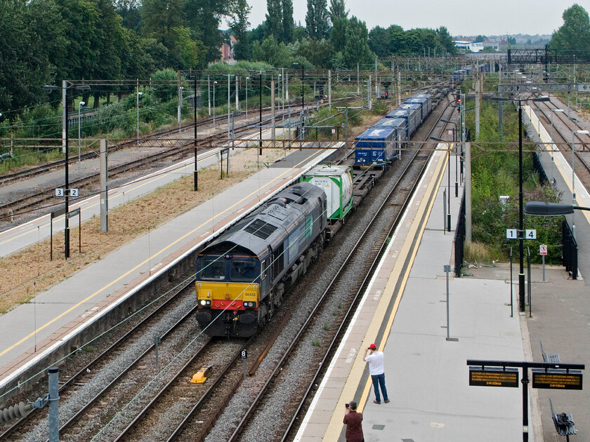 66433, 11.15 DIRFT-Tilbury FLT (4L48), Northampton station (from temporary footbridge) 
 Catching the attention of a couple of railway photographers on the platform at Northampton station 66433 brings the daily 4L48 11.15 DIRFT to Tilbury Teso Express slowly through the centre road. This new viewpoint is as a result of the new temporary station footbridge that has opened as part of the full rebuilding of the station. It certainly offers a commanding view of the lines both north (as here) and south through the station. 
 Keywords: 66433 11.15 DIRFT-Tilbury FLT 4L48 Northampton station from temporary footbridge DRS Teso Express