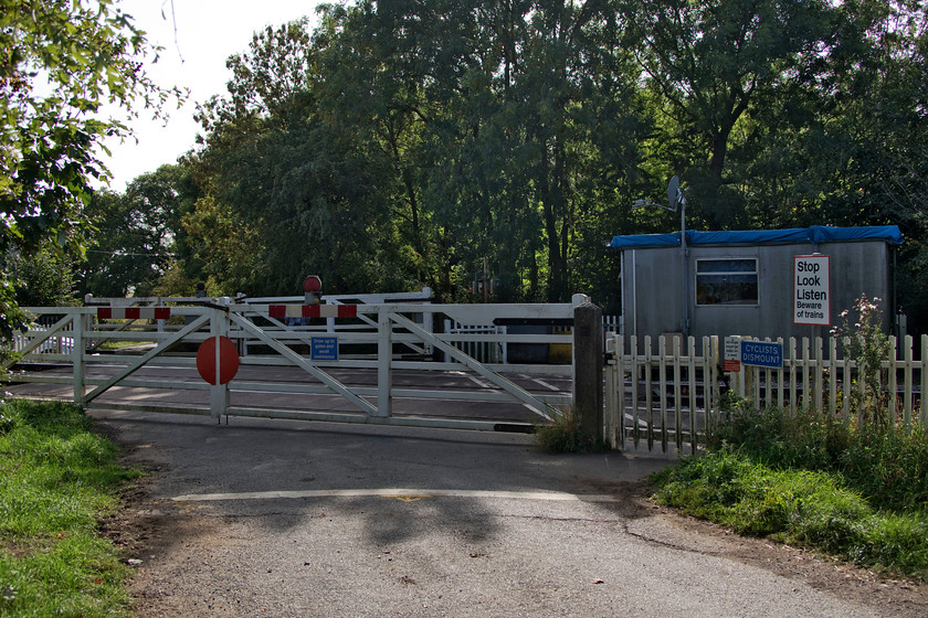 Wymondham MCG crossing 
 For a relatively busy line, and one set to carry more traffic as increasing amounts of freight is routed this way, it is unusual to find a MCG (manually controlled gates by signaler or keeper) crossing. The very quiet country lane between Wymondham and Stapleford that skirts the park of the same name crosses the line that has more trains than road vehicles hence the gates remaining closed unless a road crossing is required. The keeper's hut is a pretty dismal affair compared with a nice Victorian box that would have once been present. Notice Network Rail's attempts to keep the interior dry by covering the roof in a tarpaulin! 
 Keywords: Wymondham MCG crossing manually controlled gates by signaler or keeper