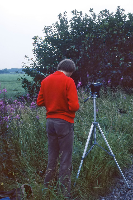 Graham, set up, Chat Moss SJ704972 
 Graham makes some adjustments to his camera whilst standing behind his trusty Velbon tripod with his Super 8mm cine camera mounted on top. He, Mike and I were waiting for the passage of 5690 'Leander' leading a BR organised special charter from Manchester to Liverpool as part of the Rocket 150 celebrations. We are standing close to the lineside on the flat expanse of Chat Moss, somewhere that one would dare not to venture on to today such is the different attitude towards trespass then and now! 
 Keywords: Graham Chat Moss SJ704972