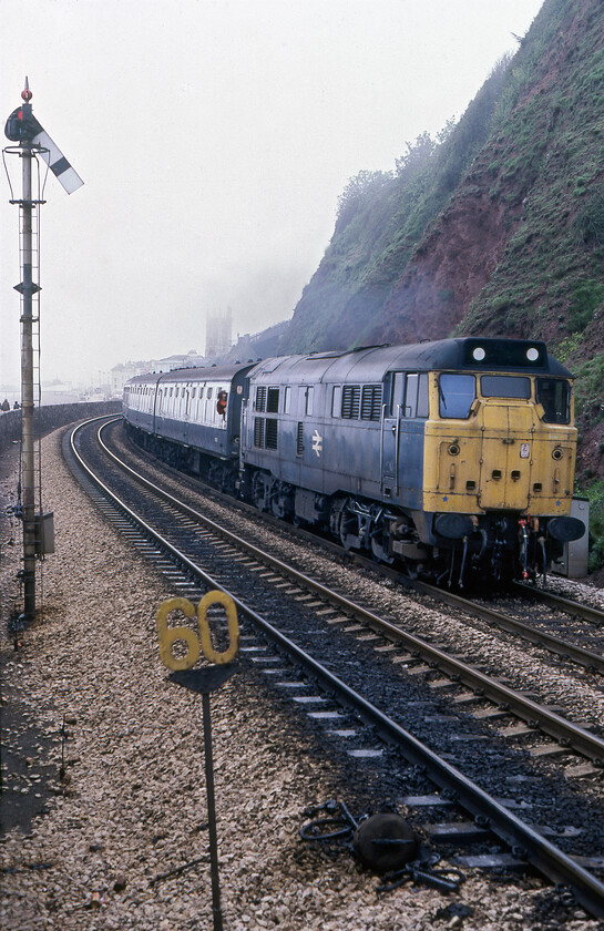 31131, unidentified up working, Teignmouth 
 With the rather ornate tower of Teignmouth's St. Michael the Archangel church largely obscured by the haar drifting in from the sea 31131 makes a racket as it accelerates away from the town's station on to the sea wall. It is passing a curiously positioned home signal some distance from the box that was located at the far end of the station. Also, notice the treddle operated track greaser on the down track and the jauntily angled sixty miles per hour speed limit sign. Unfortunately, I have not been able to identify the working but given the motive power and the stock I suggest that it may be some sort of local from Paignton; perhaps the enthusiast hanging from the first droplight may be able to advise me if he ever comes across this image! 
 Keywords: 31131 up working Teignmouth sea wall