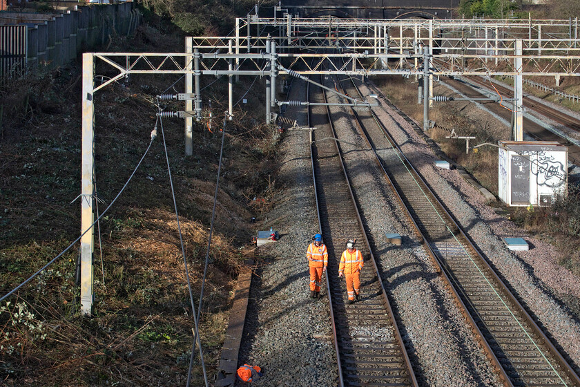 Trackworkers, site of Roade station 
 With the up and down fast lines shut for an extended period enabled by the COVID induced significant drop in passenger numbers and the consequential reduction in services, Network Rail undertook extensive work on the Weedon or 'old' route. Having been undertaking embankment clearance slightly further along the line two workers return to their access point in the January sunshine. Evidence of work undertaken a few days earlier is seen to the left of the photograph taken at the site of Roade's former station. 
 Keywords: Trackworkers site of Roade station