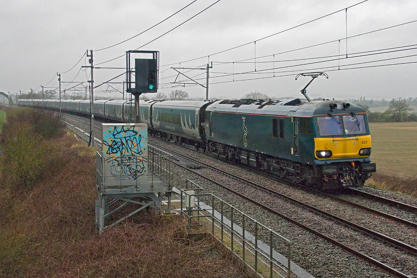 92023, CS 20.45 Inverness & 21.43 Aberdeen-London Euston (1M16, 155L), Milton crossing 
 It's February 2024 so it must be raining then! With the month shaping up to be the wettest February on record, it is absolutely pouring with rain at Milton crossing just north of Roade cutting. The reason for making the effort to take what is a pretty awful photograph is the very late running Highland Sleeper running well over two hours late. 92203 is leading the 1M16 that ran into difficulties leaving Edinburgh. At least as a passenger, you can enjoy a long lie-in if this occurs but you may well miss the start of that all-important city business meeting! 
 Keywords: 92023 20.45 Inverness 21.43 Aberdeen-London Euston 1M16 Milton crossing CS Caledonian Sleeper