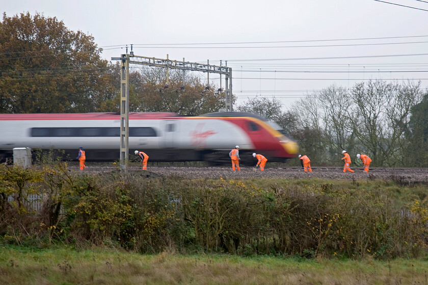 Class 390, VT 12.28 London Euston-Glasgow Central (1S67), track workers on up & down slow lines, between Roade & Ashton 
 I have used a slow shutter speed and relatively long focal length to emphasise the speed of the Virgin Pendolino passing, which appears to be, perilously close to a number of track workers who are going about their business between Roade and Ashton just south of Northampton. I had observed them for some time shovelling ballast about ready for a tamper to come and dress the stone for the track to then sit firmly in it. This is not a pleasant working environment and one that I would not want to be in especially on a Sunday morning! 
 Keywords: Class 390 12.28 London Euston-Glasgow Central 1S67 track workers on up & down slow lines, between Roade & Ashton Virgin Pendolino