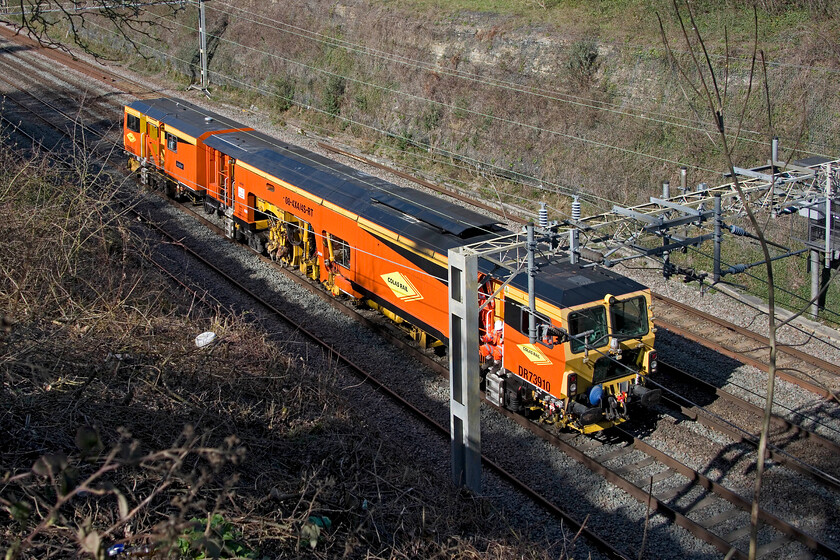 DR73910, engineering work move, Hyde Road bridge 
 With the loaded train of autoballaster wagons having reversed at less than walking pace to the work site in Roade Colas universal tamping machine DR73910 Jupiter follows at an equally slow pace. After ballasting later in the afternoon the unit will tamp the fresh stones and prepare the line for service again. Notice the orange-clad crew member sitting on the running board whilst on his mobile phone; no doubt this sitting position would not be approved in the risk assessment! 
 Keywords: DR73910 engineering work move Hyde Road bridge Colas Rail