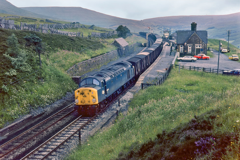 40047, down freight, Dent station 
 An absolutely superb mixed freight from a bygone era passes through the closed Dent station. I would have thought that this would be an unfitted freight given the vintage of the wagons in the consist, but close examination of the rear of the train does not indicate a brake van. 40047 is leading the train on the final part of the more gentle climb of the fabled 'Long Drag' from the south with just a few miles to the summit at Ais Gill. I had haulage behind 40047 in March 1984 when it was one of the locomotives working The Conway Crusader railtour along with 50018 'Resolution'. The locomotive was withdrawn in late 1984 but lingered on eventually being cut up by January 1986 at Doncaster. Notice the cars parked in the former station car park, of particular interest, is a Fiat 124 (or, is it an early Lada version?) behind the Rover SD1, the inevitable Mini and a Scimitar GTE parked by the fence at the rear. 
 Keywords: 40047 down freight Dent station