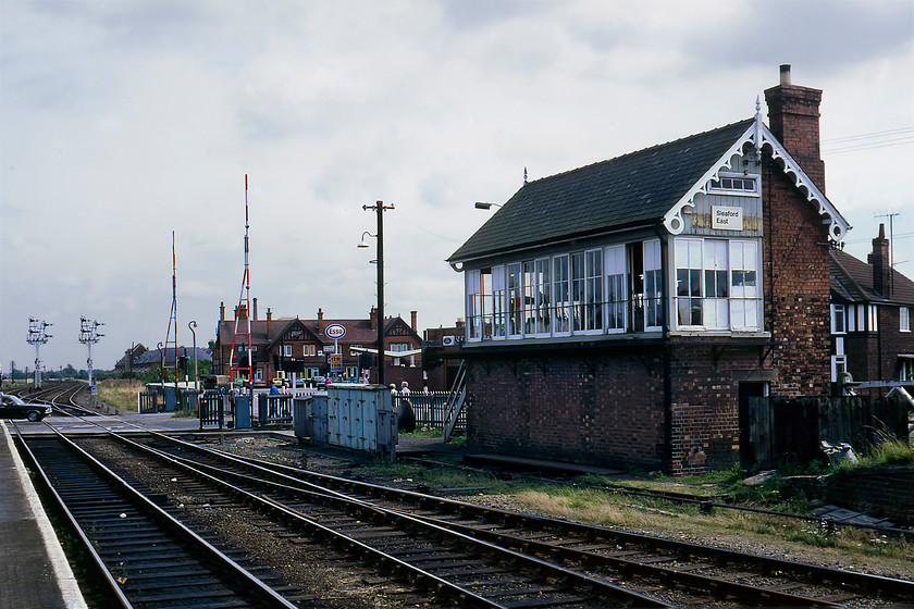 Sleaford East signal box (GN, 1882) 
 Sleaford East signal box is located (as it still is due its Grade II listing) between the eastern end of the station and the level crossing with Grantham Road. It's an 1892 Great Northern building that has been somewhat spoilt by the crass application of one of BR's corporate image signs. In this view notice the 'Coke bottle' Ford Cortina Mk. III crossing the level crossing. The large building behind the Esso petrol station is still in existence and looks exactly the same but the Ford dealership has now gone to be replaced by an Aldi supermarket. The superb pair of twin doll brackets on slotted posts round off a really interesting scene in this small Fenland town. 
 Keywords: Sleaford East signal box Great Eastern Railway