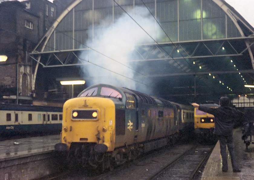 55015, 16.05 London Kings Cross-Leeds & Harrogate (1L26) & 47459, unidentified down working, London Kings Cross station 
 With its characteristic plume if smoke emerging from the exhaust, 55015 'Tulyar' leaves King's Cross with the 1L26 16.05 to Leeds and Harrogate. My school friend and spotting companion, Guy is seen holding his microphone out to capture the 'Napier Magic' of the departure although looking at the smoke emerging from one side only, could it be that 55015 was only running on one engine as was fairly common? Behind Guy's outstretched arm, 47459 waits with an unidentified down working. At the time of writing in 2018, Tulyar is in the final phase of its long and protracted rebuild by the amazing team at the Deltic Preservation Society (of which I am life member) at Barrow Hill. So, could it be that the mighty 55015 could again be seen leaving King's Cross for the north sometime soon? 
 Keywords: 55015 16.05 London King's Cross-Leeds Harrogate 1L26 47459 unidentified down working London Kings Cross station