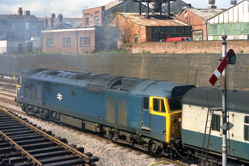 50003, unidentified down working, Newbury station 
 The up starter has been pulled off and 50003 makes a noisy and smoky departure from Newbury station. This view of the 50 clearly shows that its not refurbished (The programme had not yet started with 50006 being the first completed in 1979) as the air vents in the roof cut-out are still exposed. During the refurbishment process, this large space was covered up with a new section of roof panel and the whole filtration system heavily revised. Unfortunately, this had the effect of removing the sucking of air sound that gave the 50s their 'Hoover' nickname. Notice the stack of track panels in the foreground that had been removed from the west facing sidings and bay platform. 
 Keywords: 50003 down working Newbury station