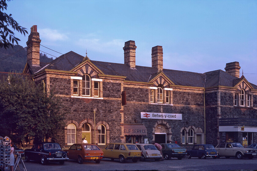 Frontage, Betws-y-coed station 
 After a superb day surveying the North Wales coastal route Graham and I headed for home in the Auston 1100, indeed the car is seen fourth from the left in this photograph. We must have stopped at Betws-y-coed for a comfort break as we had already taken a brew-up at the top of the Nant Ffrancon valley at the foot of Tryfan. The large station building looks grand in the evening light as does the superb lineup of cars the rarest today is probably the yellow Fiat 128 estate next to our 1100. The car park has largely gone from the front of the station today with it being pedestrianised. 
 Keywords: Frontage Betws-y-coed station