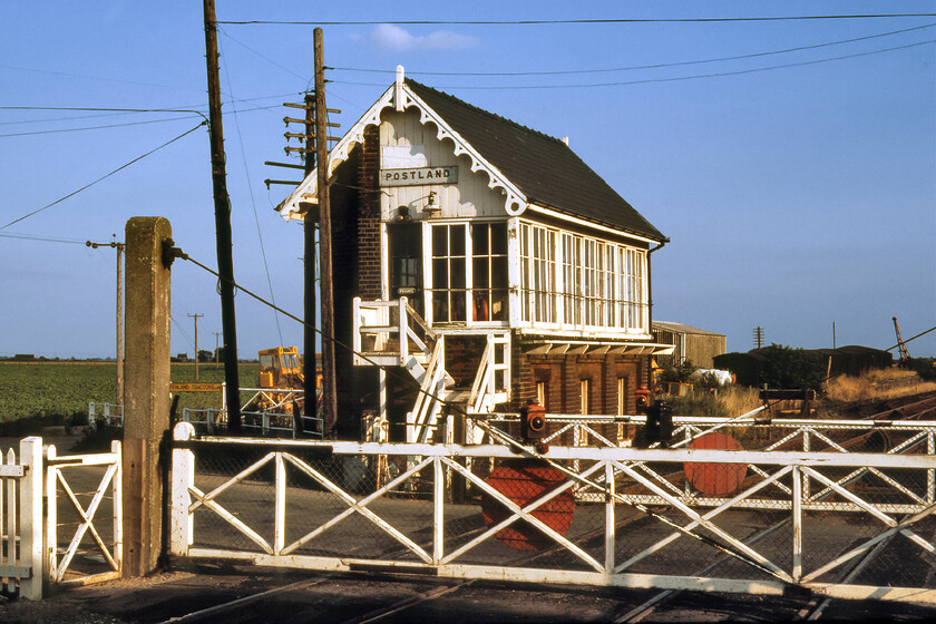 Postland signal box & crossing gates (GN, 1872) 
 Postland signal box looks to be in superb condition on this warm and pleasant sunny August Sunday afternoon back in 1981. Whilst the box still stands today, it is not in quite such a good way in fact it is almost hidden behind a cloak of ivy with the roof falling in, see.. https://www.ontheupfast.com/p/21936chg/30044473949/x15-former-postland-signal-box-gn . The ornate box with its characteristic sculptured barge boarding was (is!) a Great Northern design built in 1882. To take this photograph, including the superb crossing gates with their oil lamps, I was standing on the down platform of the former station that itself was closed in 1961. Fenland Tractors, seen behind the box, is still in operation today and indeed, a Google search for them reveals the rear of the box on the search page. 
 Keywords: Postland signal box & crossing gates Great Northern