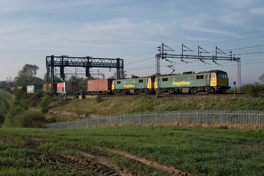 86632 & 86612, 02.50 Felixstowe North-Garston (4M45, 63E), between Roade & Ashton 
 The 02.50 Felixstowe to Garston Freightliner passes between Roade and Ashton led by 86632 and 86612. This image shows a generational change between WCML overhead line structures. The leading ac electric is passing under one of the original latticed stanchions installed during the 1960s electrification. Above the leading wagons is a gargantuan and chronically over-engineered structure spanning the entire four lines that supports just two up colour lights that dates from the early 2000s. 
 Keywords: 86632 86612 02.50 Felixstowe North-Garston 4M45 between Roade & Ashton Freightliner AL6 AC electric