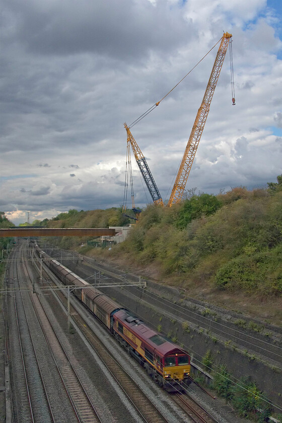 66013, 14.41 DIRFT-Dollands Moor (6O67, 3E), Roade cutting 
 EWS liveried 66013 leads the 14.41 Daventry IRFT to Dollands Moor empty water train through Roade cutting. Towering above the cutting is a substantial crane that had installed the girders seen spanning the lines. These girders will support Roade's new bypass that will circumvent the village to the western side; something that the locals have been desperate for many years. However, being funded by a private company rather than by the highways or local authorities tells a further tail. As a planning sweetener it was agreed that it should be privatly funded if planning was given for the nearby and absolutely huge rail connected SERGO Logistics Park; some locals are concerned that the development is a price too far. 
 Keywords: 66013 14.41 DIRFT-Dollands Moor 6O67 Roade cutting EWS Class 66 water train