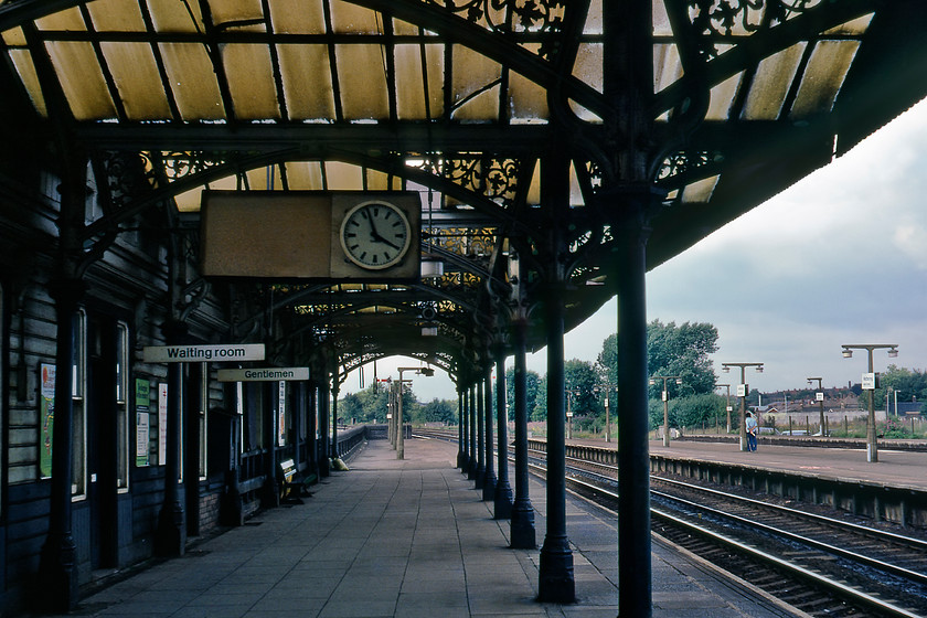 Down fast platform, looking north, Kettering station 
 Platform four of Kettering station (the down fast) is seen looking north. This platform was constructed in 1879 along with the island platform (two and three) to match the original 1857 station building designed by the esteemed architect C H Driver. In this photograph, the single-storey weather-boarded waiting rooms and canopies, with cast-iron columns and spandrels are clearly seen. The condition of the glass is clear to see here, as well as being badly stained it is also cracked. BR in its cost-saving wisdom wanted to remove the canopies and glass to be replaced by plastic sheeting. Thankfully, Kettering Civic Society got to hear of their plans and created a fuss meaning that the plans were withdrawn. Eventually, the whole structure was comprehensively refurbished by Railtrack in 2000. 
 Keywords: Down fast platform looking north Kettering station Midland Railway