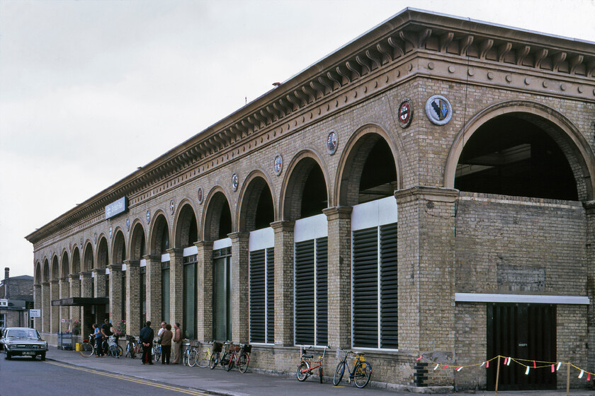 Frontage, Cambridge station 
 I am sure that when Cambridge station opened in 1845 its long classical faade and porte-cochre (crudely infilled during the 20th century as seen here) it seemed a large and substantial building. Today it is very similar to this 1981 view apart from the removal of the slats to be replaced by glazing but the building is now dwarfed by the huge developments around it. Designed by Sancton Wood and Francis Thompson the station is now Grade II listed. Notice the bikes, including a Raleigh Chopper leaning against the front of the station, being holiday time I suspect that they did not belong to students. Also, spot the 1973 Peterborough registered brown Audi 80 at the front of the station that was last on the road in early 1989. Of note are the coats of arms that adorn the frontage located between the arches. Carved and painted in relief twenty-nine were installed when the station was built. They are the coats of arms of various local noteworthy people, the colleges and one of the City of Cambridge itself. 
 Keywords: Frontage Cambridge station