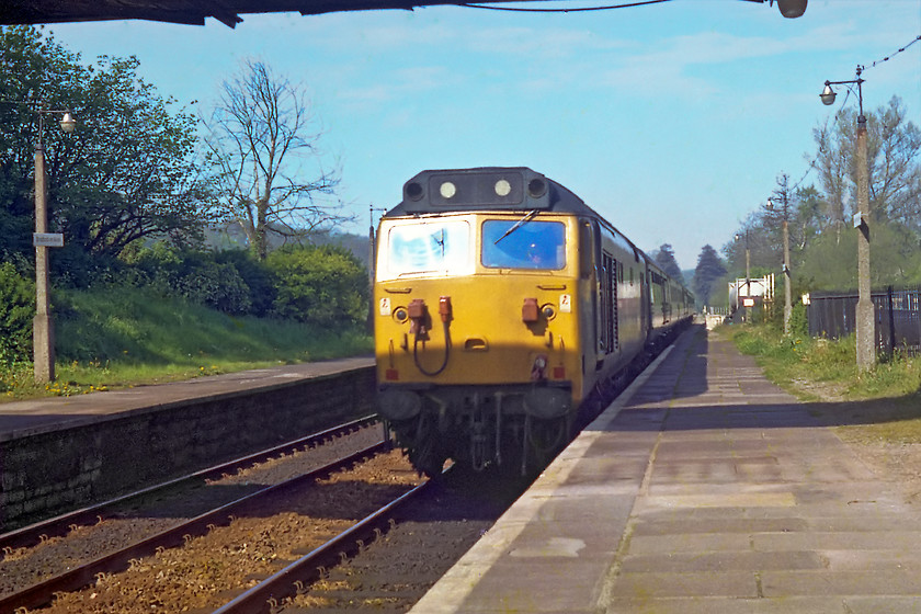 50027, unidentified up diversion, Bradford-on-Avon station 
 The up platform at Bradford-on-Avon was controlled by a sole colour light (BJ102). This was, in fact, a very distant-distant from Bradford Junction signal box some three miles away. If a through train was due, BJ102 would show green and remain so for some length of time prior to the arrival of the train. I believe that the green was given after the train had passed over Bathampton Junction but I could be wrong? Us spotters would all get very excited if this happened as it heralded the arrival of something unusual. Our eyes would be peeled looking up the one mile or so of straight track west of Bradford-on-Avon in anticipation of what was coming! On this particular morning, this event heralded the passing of an up diversion to London Paddington headed by 50027. I had to go for these more head-on shots to avoid the plague of motion blur as the Exa camera had a maximum shutter speed of 1/150 sec. However, my timing was not always spot-on as demonstrated by the shadow cast by the footbridge! In recent years, rampant tree growth has altered this viewpoint considerably, see..... https://www.ontheupfast.com/p/21936chg/29201867204/x150238-13-10-weymouth-gloucester 
 Keywords: 50027 Bradford-on-Avon station Paddington