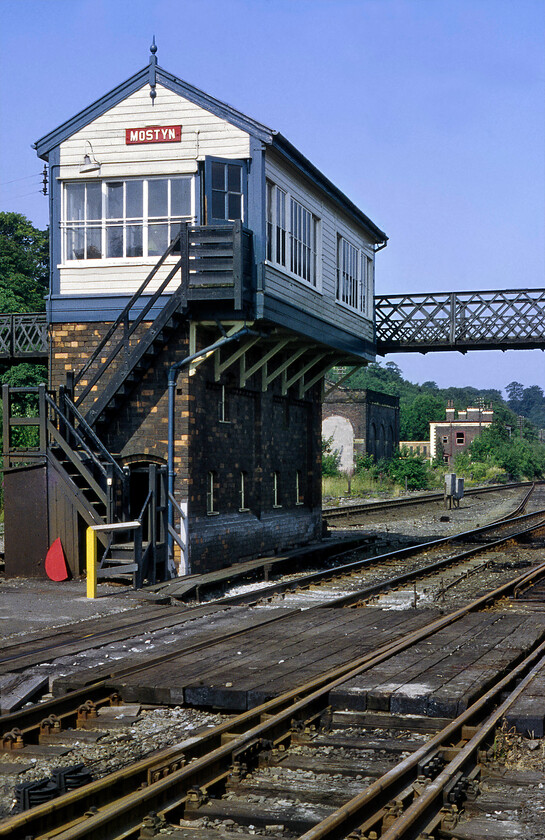 Mostyn signal box (LNW, 1902) 
 When the North Wales coastal route was largely quadrupled by the L&NWR there was plenty of room along much of its length. However, there were some tight spots such as here at Mostyn where there was a busy harbour and a number of sidings that serviced it. The running lines had to be closer together so the LNWR had to construct a narrower than normal signal box that required a cantilevered operating floor. Back in 1981 the box was still very much in use with a footbridge (now removed) crossing the lines behind in this view with the closed station and goods shed further beyond that. To take this photograph I was standing on a level crossing that afforded access to the harbour. 
 Keywords: Mostyn signal box LNWR London & North Western Railway