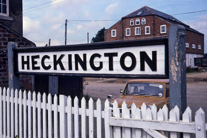 Running in board, Heckington station 
 A superb timber running in board at Heckington station. This particular example on the eastbound platform (towards Skegness) still exists today. Notice 'UAM' the orange Mini parked in the station car park behind the sign. Close examination of the car reveals that Graham was happy with two rearview mirrors both on the offside but none on the near side. However, he did have one of those extra-wide rear view mirrors that attached over the original with springs that were so popular in the 1970s! 
 Keywords: Running in board, Heckington station