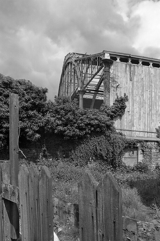 Trainshed, Bath Green Park station, from Midland Bridge Road 
 Taken from Bath's Midland Bridge Road the impressive arch of Green Park's trainshed stands out well in the spring sunshine against a dark sky. I really like this photograph that has utilised the excellent depth of field afforded by my Pentax's superb prime lens by closing the aperture down as far as I dare go. This has meant that as well as having the trainshed in sharp focus so is the superb and extremely weathered boundary fence in the foreground. This shot can be almost exactly recreated today but the fence has no long gone as has the derelict building out of site immediately to my left. 
 Keywords: Trainshed Bath Green Park station from Midland Bridge Road