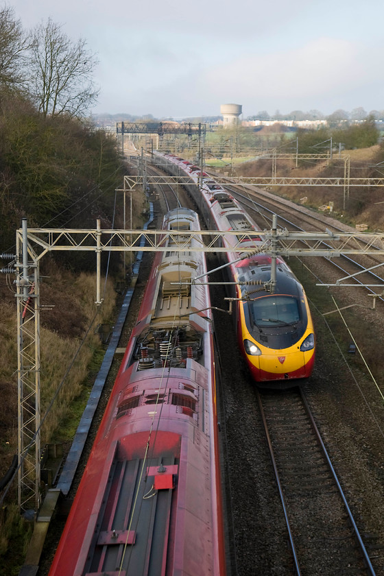 Class 390s, VT 12.28 London Euston-Glasgow Central (1S67) & 10.58 Preston-London Euston (1A16), Victoria bridge 
 At a passing speed in the region of two hundred miles per hour, two class 390s cross at Victoria bridge between Northampton and Milton Keynes. The down service to the left is the 12.28 Euston to Glasgow Central whilst the up train is the 10.58 Preston to Euston. In the background is one of Roade's iconic water towers with them being located in the village with it being the highest part of this area of Northamptonshire. 
 Keywords: Class 390s 12.28 London Euston-Glasgow Central 1S67 10.58 Preston-London Euston 1A16 Victoria bridge