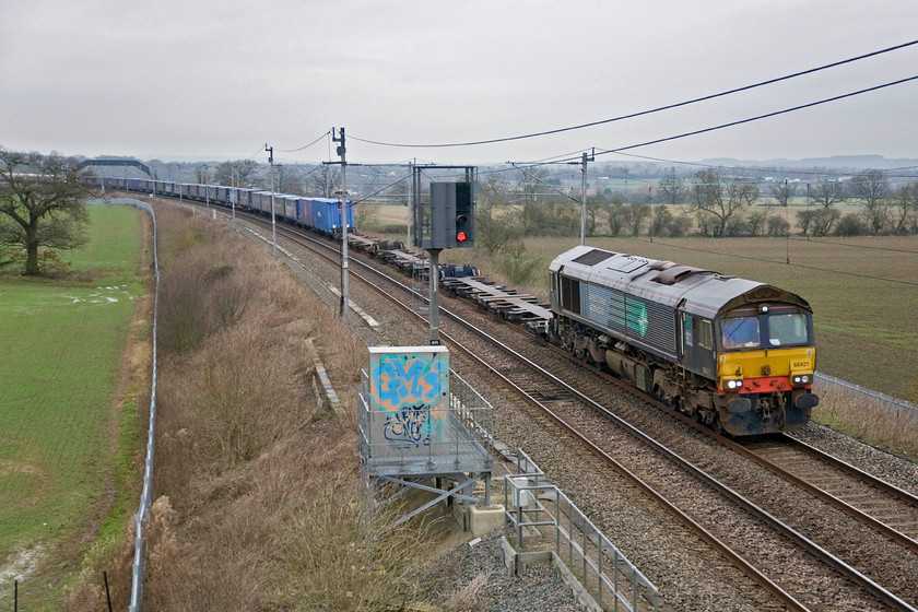 66421, 11.01 Daventry-Purfleet (4L98), Milton crossing 
 Wearing its DRS compass livery 66421 works the Sunday 4L98 Tesco Express (as it has been dubbed) past Milton Crossing a short distance north of Roade. This working leaves Daventry for Purfleet today at 11.01 but usually a bit later. It has left earlier than normal as it initially had to head north to Rugby for the 66 to then run round and bring it south on the 'mainline' via Weedon avoiding Northampton due to engineering works. I was hoping for some sunny weather at this classic location but the bright weather of earlier had been replaced by cloud and dullness with it now spitting with rain; all the best plans and that! 
 Keywords: 66421 11.01 Daventry-Purfleet 4L98 Milton crossing DRS Direct Rail Services Tesco Express