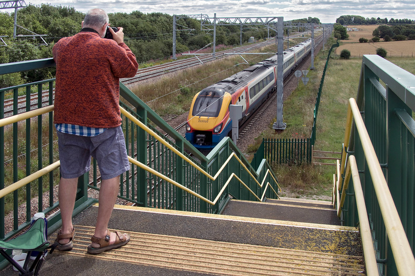 Andy & 220020, EM 11.31 London St. Pancras-Sheffield (1F27, RT), Irchester SP927667 
 Andy takes his photograph of 222020 as it passes Irchester just south of Wellingborough. The EMR Meridian is working the 11.31 St. Pancras to Sheffield service. We are standing on the massively over-engineered footbridge that spans the four tracks of the MML that replaced a very rickety structure a couple of years ago. 
 Keywords: Andy 220020, EM 11.31 London St. Pancras-Sheffield 1F27 Irchester SP927667 EMR East Midlands Railway