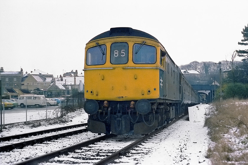 33027, 08.56 Portsmouth Harbour-Bristol Temple Meads, Bradford-on-Avon station 
 Complete with a set of mini snow ploughs 33027 is sporting the wrong Southern Region reporting code heading the 08.56 Portsmouth Harbour to Bristol Temple Meads working. 85 was a Portsmouth to Southampton/Romsey slow working, it should have been 89 for the working pictured. The driver peers back waiting for the right away from Bradford-on-Avon station, next stop would be Bath Spa. Notice the collection of 1970s cars in the station car park dominated by the VW camper. No fewer than five of the cars seen are Fords of various types. 
 Keywords: 33027 08.56 Portsmouth Harbour-Bristol Temple Meads Bradford-on-Avon station