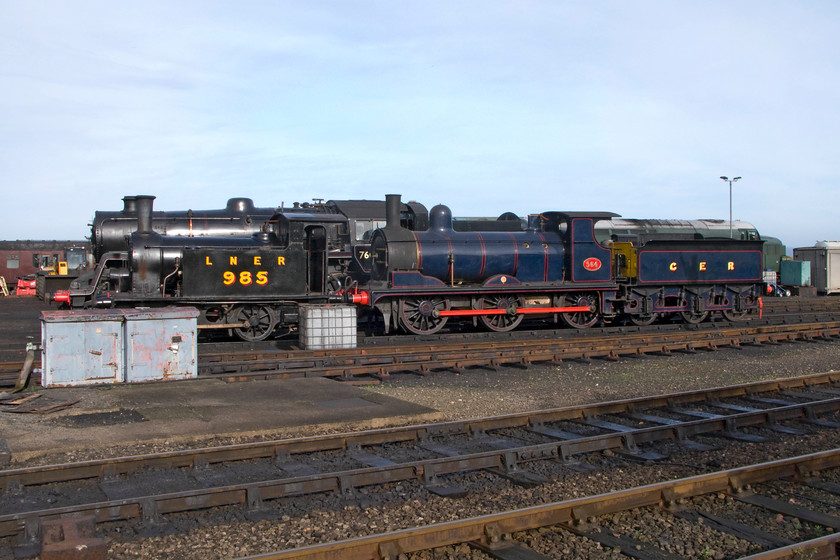 985, 76084, 564 & D6732, stabled, Weybourne yard 
 An interesting and eclectic collection of motive power is seen in Weybourne yard. Y7 number 985 was built in 1923 by the LNER to an 1888 NER design for work on short coal trains primarily shunting them around collieries and docks of the northeast. For ease of sighting when backing up to wagons notice 985 has no rear bunker having small ones located within the cab itself. 985 was in use until withdrawal came in 1964 after many years of industrial use. Dominating the view is former GER Y14 number 564. It is quite at home here in Norfolk as it spent its entire fifty-one years of operation in East Anglia being based at many different depots. It was withdrawn in 1963 when steam in East Anglia was eliminated whilst acting as a Liverpool Street station pilot. At this point, it had outlived many much more modern designs. Behind the two vintage locomotives is the black hulk of the far more modern 4MT 76084. This Mogul had a working life of a mere ten years from 1957 to 1967. It was constructed with a forty-year operating life span expected by Horwich works. This would have meant that withdrawal would have been anticipated by 1997 only four years before Voyagers arrived on our railway! Hiding behind the three steam locomotives is Type 3 D6732. This dates from 1962 and is in regular operation on the North Norfolk Railway. As 37032 it was variously based around the northeast spending many years at Thornaby. 
 Keywords: 985 76084 564 D6732 Weybourne yard