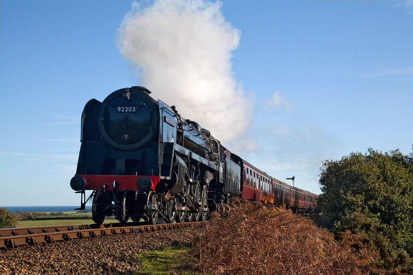 92203, 10.45 Sheringham-Holt, Kelling Bank 
 Sun, sea and steam....what more can be asked for? The 10.45 Sheringham to Holt working is seen climbing Kelling Bank with 92203 'Black Prince' putting on a good show. The bank is pretty steep with it rising straight away from Weybourne station about a mile from this spot. Some locomotives struggle to get going but the 9F makes easy work of the task and takes on the climb with relative ease. 
 Keywords: 92203 10.45 Sheringham-Holt Kelling Bank