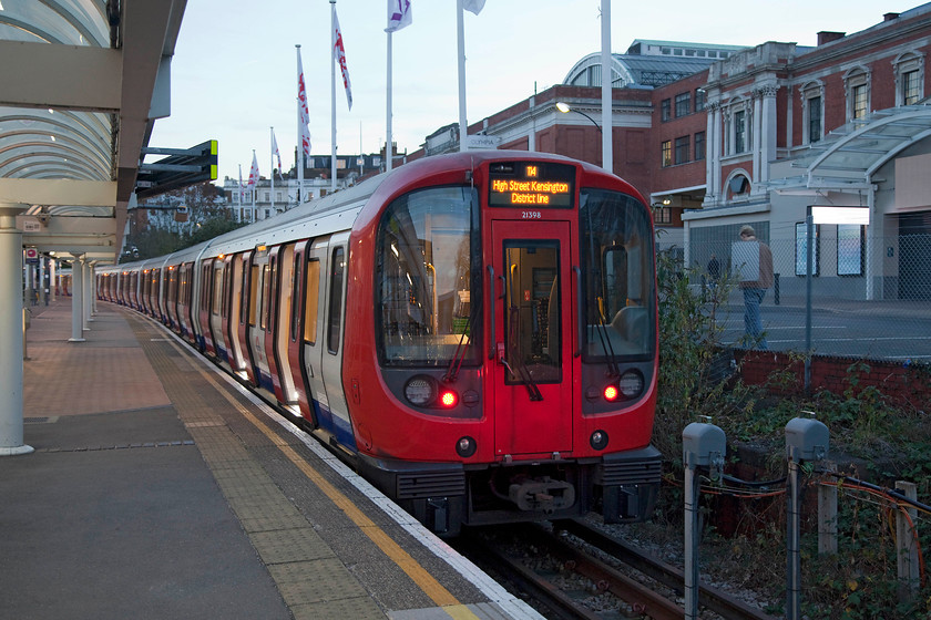 21398, Kensington Olympia-High Street Kensington District working, Kensington Olympia station 
 With the Kensington Olympia Exhibition Centre dominating the background, TFL 21398 waits at the adjacent underground station platform. This unit is one of the new S7 trains that are being introduced at the time of writing. Whilst they do not have the character of the old stock, they represent a huge step forward for London travellers, the main advantage is that thy are air-conditioned. 
 Keywords: 21398 Kensington Olympia-High Street Kensington District working Kensington Olympia station