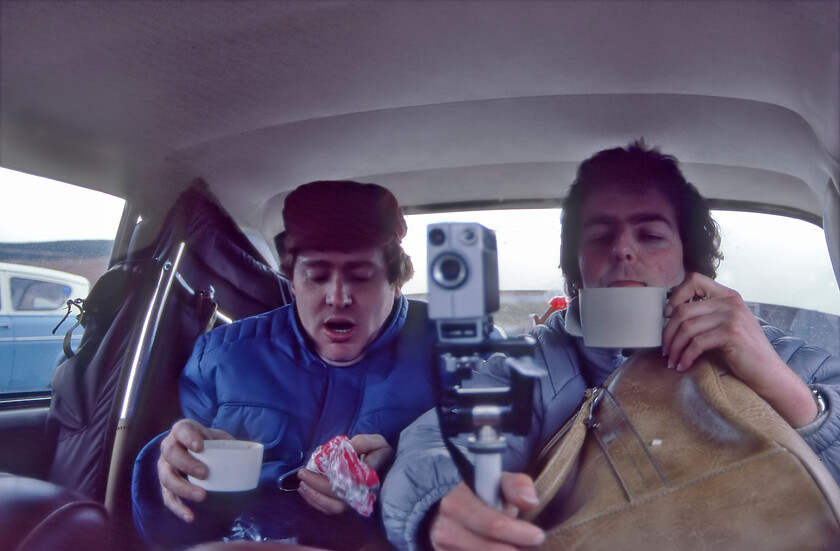 Paul & David, between spots, UVJ 129J 
 When the chase is on there is no time to stop and have a brew! Paul and David attempt a mouthful of tea from their flasks in the back of the Austin 1100. Notice the appropriate era related apparel as worn by any self-respecting gricer along with their accessories in the form of rucksacks, bags and a Super8 cine camera wisely mounted on a Velbon tripod. Also, notice the dual coloured Ford Anglia Super parked next to the 1100, perhaps it was owned by a fellow gricer as there were many about on this day! 
 Keywords: Paul David between spots UVJ 129J Austin 1100 gricing