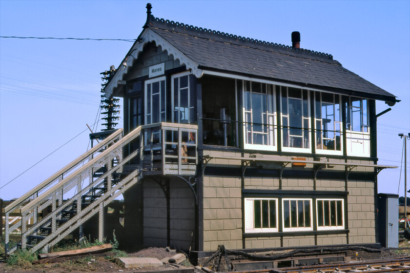 Manea signal box (GE, 1883) 
 Manea signal box is seen basking in the afternoon sunshine with the signalman having opened his huge original window with its distinctive and unusual glazing pattern. The box is an 1883 Great Eastern Type 3 design common to many in this area. Notice the steps that appear to have been made of some sort of industrial gauge Dexion? However, it still maintains it elegant bargboarding, decorative ridge tiles and one finial. In 1992, the box became the fringe box to Cambridge PSB and, at the time of writing in 2023, it is still in use but looks very different to this 1981 view. 
 Keywords: Manea signal box GE Great Eastern