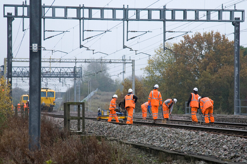 Track workers on up & down slow lines, Ashton Roade bridge 
 Whilst two tampers wait track workers manually shovel ballast on an area of track between Roade and Ashton. There were a number of worksites on this murky Sunday morning in the vicinity all employed undertaking the same type of work. 
 Keywords: Track workers on up down slow lines Ashton Roade bridge Tampers