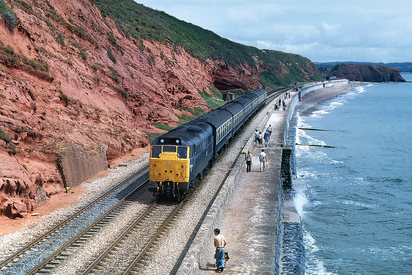 31256, down ECS, Dawlish SX971773 
 With the famous Langstone Rock in the distance, 31256 brings an empty coaching stock working along the sea wall composed of a short rake of Mk.1s. The characteristic red Dawlish Sandstone that has caused so many problems with erosion can be seen clearly in the cliff on the left. The sun has decided to come out and as a result, the visitor walking the pushchair along the sea wall has his shirt off but has kept his very 1970's flared jeans on! 
 Keywords: 31256 ECS Dawlish SX971773