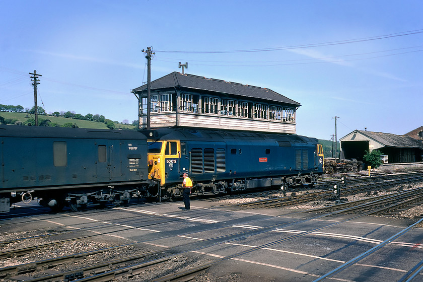 50012, 10.05 Paignton-London Paddington, Exeter St. David`s station 
 50012 'Benbow' passes Exeter Middle signal box over the level crossing that is being watched over by the crossing keeper. On a summer Saturday, the crossing keeper would be kept busy ensuring that he struck a balance between keeping everything safe for all users but at the same time avoiding hefty build-ups of road traffic on the approaches to the level crossing, especially on the busy Cowley Bridge road. He was also permitted to allow pedestrians to cross under his directions when the barriers were lowered through side gates. This was the time when us humans had something called common sense! 50012, leading the 10.05 Paignton to Paddington, would stay in service until November of this year before being sent to Doncaster to be comprehensively re-built and emerge on 05.05.81. 
 Keywords: 50012 10.05 Paignton-London Paddington Exeter St. David`s station Benbow