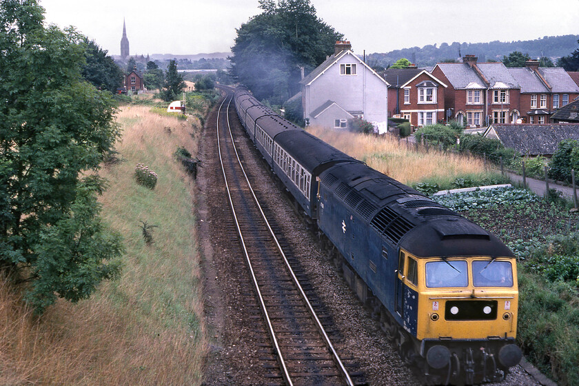 47089, 15.10 Portsmouth Harbour-Cardiff Central (1V30), Skew bridge, Bemerton 
 Having seen 47089 'Amazon' work south earlier in the day, see..... https://www.ontheupfast.com/p/21936chg/30035687461/x47089-08-15-cardiff-central-portsmouth it is captured on its balancing working, the 15.10 Portsmouth Harbour to Bristol Temple Meads. Picking up speed after leaving Salisbury the train is seen about to pass under the A36 road's Skew bridge in the suburb of Bemerton. Notice the railway land, unprotected from the running lines, to the right of the photograph that has been acquired for use as an allotment. Activities such as this would have been unofficial but very much tolerated by the railway authorities at this time; how different to today! 
 Keywords: 47089 Amazon 15.10 Portsmouth Harbour-Cardiff Central 1V30 Skew bridge, Bemerton