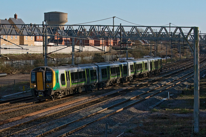 350122, LN 13.31 Bletchley carriage sidings-Northampton ecs (5N55, RT), site of Roade station 
 The village of Roade is the home to two water towers but neither are in use now. The one in this image provided huge amounts of water to the former Pianoforte factory that occupied the site of the new houses. The concrete structure is currently undergoing conversion into a private residence. See.... https://www.freehausdesign.com/ashton-road-water-tower 350122 hurries past the new housing estate to the south of the village with the 13.31 Bletchley to Northampton ecs working. 
 Keywords: 350122 13.31 Bletchley carriage sidings-Northampton 5N55 site of Roade station