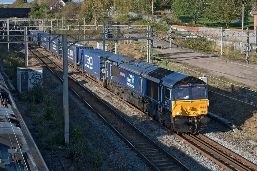 66428, 13.51 DIRFT-Tilbury (4L48, 12L), site of Roade station 
 As an Avanti West Coast Pendolino speeds southwards 66428 'Carlisle Eden Mind' makes more sedate progress leading the 4L48 13.51 Daventry (DIRFT) to Tilbury 'Tesco Express'. The two trains are passing through Roade at the site of the former station. The gap between the fast and slow lines was once occupied by the central island platform with the rather basic station building in an elevated position on the top of the bank above the third box of the freight with the two linked by a rather rickety timber footbridge. 
 Keywords: 66428 13.51 DIRFT-Tilbury 4L48 site of Roade station DRS Direct Rail Services Carlisle Eden Mind