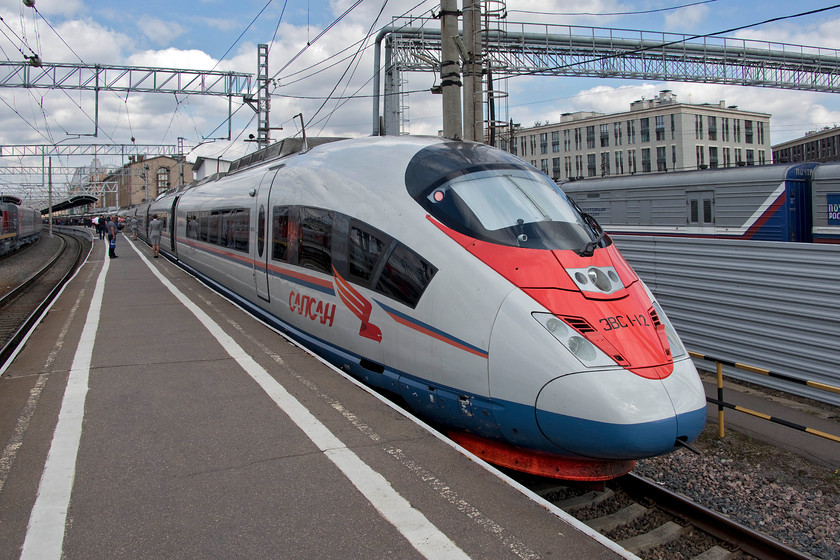 EVS1-12, 13.00 St. Petersburg-Moscow (765A), St. Petersburg Moskovsky station 
 In a break from the usual grey livery applied to most RZD rolling stock, their headline trains, the EVS1 Sapsans are painted white. Looking purposeful sitting at St. Petersburg Moskovsky station EVS1-12, with another set attached to the rear, waits to leave with the 13.00 to Moscow. We took this train to the Russian capital city. Notice the grey suited staff standing by every coach door. Their job was to greet and admit passengers after scanning their passports or Russian ID cards and directing them to their seats. They also then travelled on the train in that carriage meaning a total of twenty RZD staff were on the train composed of two ten car sets; an unimaginable situation when compared to the UK! The Sapsans are a widened version of DB's ICE train, see.... https://www.ontheupfast.com/v/photos/21936chg/24262781204/x406054-13-29-frankfurt-hbf-amsterdam that takes full advantage of the generous Russian railway loading gauge. This wider body was noticeable inside the train with very generous seating and large gangways. In addition, the seats were very comfortable, Hitachi and other manufactures of UK trains need to take note! 
 Keywords: EVS1-12 13.00 St. Petersburg-Moscow 765A St. Petersburg Moskovsky station