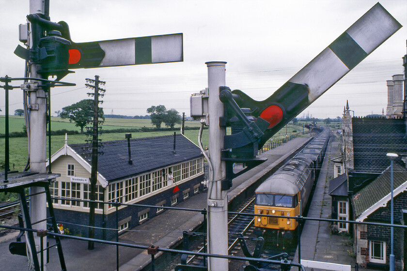 Class 56, up empty hoppers, Brocklesby station 
 The waving of a red flag by, in this case, the signalmen in Brocklesby signal box is an urgent request for a train to stop. As the up empty hopper train hauled by the Class 56 has the road and has passed the box I suspect that the signalman was not requesting this to stop but that he was flagging down a down train that would have been coming into view behind me. Either way, my contemporary notes do not make clear what I had observed apart from a Class 47 leading a freight towards Scunthorpe. In this wide-angled view taken from the B1211 road bridge, the mock Tudor Gothic-styled station can be seen to the right with the unusually placed Great Central signal box on the island platform. This scene is essentially the same today apart from the absence of the bracket signal and the rest of the signalling equipment. Additionally, whilst this particular Class 56 has probably been scrapped a small number are still in use on the network. 
 Keywords: Class 56 up empty hoppers Brocklesby station