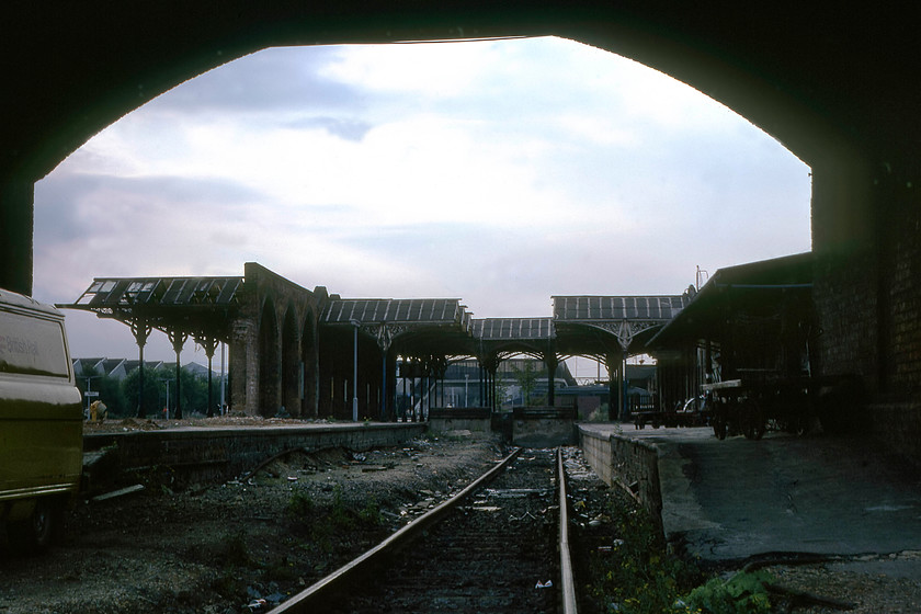 Bedford Midland station (Closed October 1978) 
 An evocative view of the half-demolished and now derelict Bedford Midland station. The photograph is framed by the arch of Ford End Road bridge that spanned the lines south of the station. Through the remains of the wrought iron roof work of the former station, the new (and present day) station can be seen complete with the ugly new footbridge spanning the recently aligned MML tracks. The new station had opened some ten months prior to this image being taken. Notice the British Rail Commer PB van to the left, there were thousands of these vans back in the 1970s and 1980s. Today, it's white Transits with Network Rail emblazoned on the sides. This image can be replicated today with the same archway framing the top, but the view would be of a huge carpark serving the many commuters using the station. 
 Keywords: Bedford Midland station