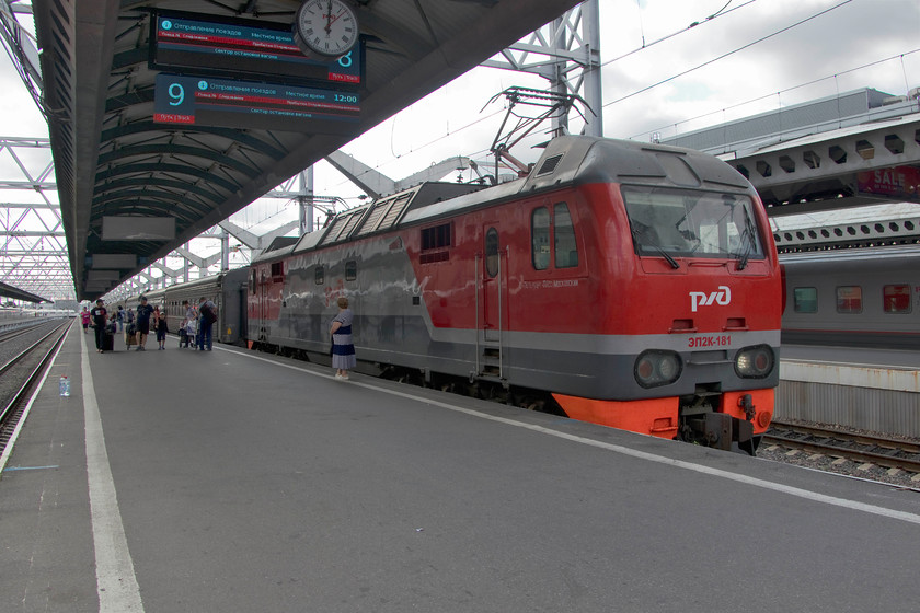 EP2K-181, stabled, St. Petersburg Moskovski station 
 The lady standing next to EP2K-181 at St. Petersburg Moskovsky station makes you realise how large these locomotives are. This new design was launched in 2005 and, testament to its success, a further large order was placed in 2016. With some 6,400hp they are powerful locomotives that are designed to haul passenger trains all over the RZD network at speeds of up to 100mph. This particular example had arrived earlier with an unidentified working and was waiting for one of the station pilots to remove the stock from behind it. 
 Keywords: EP2K-181 St. Petersburg Moskovsky station