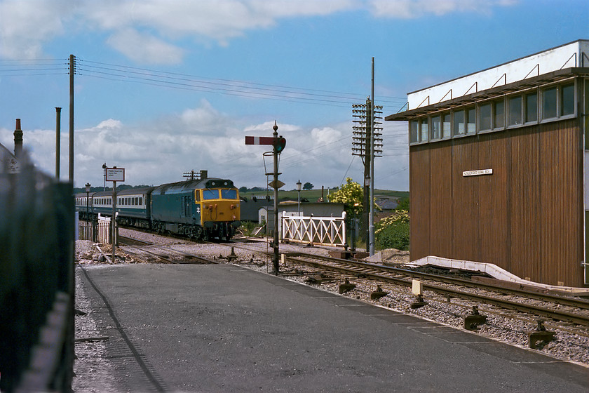 50031, unidentified up working, Hungerford station 
 50031 races through Hungerford station with an unidentified up working. It is about to pass Hungerford signal box that replaced the original Great Western structure in 1971. This followed the derailment of a freight train that partially destroyed the old box that was situated just on the other side of the crossing gates seen in this image. Examination of 50031's bodyside reveals that it has been prepared, by the moving of the BR double arrow from the centre to the cab, in order for it to be named 'Hood' two days after this picture was take that may also account for its smart appearance. 
 Keywords: 50031 up working Hungerford station