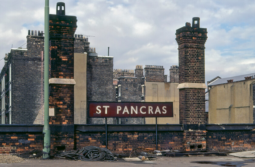 Enamel, London St. Pancras station 
 Yet another large London Midland enamel at St. Pancras station. This huge running in board was located at the extreme country end of the station on the eastern side. Notice the general detritus laying about on the platform not exactly providing the expected welcome for travellers! I love the chimney stacks on the buildings in the background with their vast numbers of pots. One can only wonder what the interiors of these buildings would have been like at this time with them being divided into many poor-quality flats and bedsits. All in this view has now been swept away with a council office and leisure centre now dominating this view.

UPDATE..... the last sentence that I wrote here is actually incorrect! When visiting St. Pancras in the autumn of 2022 I happened to spot some of the chimneys featured in this photograph. They appear to be the two sets located above the last few letters of the word Pancras with one partially obscured by the right-hand taller chimney, see..... https://www.ontheupfast.com/p/21936chg/30033204428/x2-chimneys-london-st-pancras-station 
 Keywords: Enamel London St. Pancras station