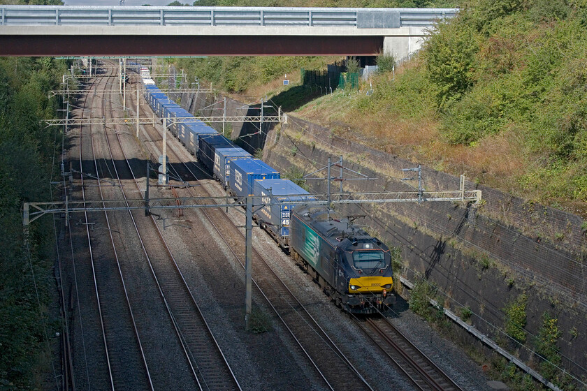 88001, 12.41 DIRFT-Tilbury Container Terminal (4L48, 4L), Roade cutting 
 88001 'Revolution' roars southbound through Roade cutting leading the seven-day-per-week 4L48 Tesco express. The speed which the electric Class 88 lift their trains up the steady incline from Northampton compared to a Class 66 is stark making full use of their ample five thousand four hundred horsepower. Just entering some shade caused by a well-timed cloud the train is the 12.41 Daventry (DIRFT) to Tilbury service. 
 Keywords: 88001 12.41 DIRFT-Tilbury Container Terminal 4L48 Roade cutting Tesco Express DRS Revolution