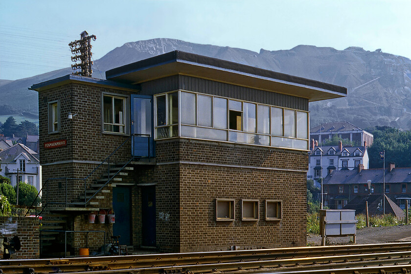 Penmaenmawr signal box (BR, 1952) 
 Penmaenmawr signal box is seen in the afternoon sunshine located at the eastern end of the station platform the ramp of which can be seen to the extreme right. I am not too sure as to why I trespassed off the end of the platform to take this view into the sun but it may well have been to include the dramatic granite quarry that dominates the landscape around this North Wales town. The box is a British Railways (London Midland) Type 14 structure dating from 1952. It replaced a poorly located L&NWR box at the western end of the station following a multi-fatal collision on the night of 27.08.50 dubbed the Irish Mail Train Disaster. In the enquiry, it was found that the light engine locomotive awaiting access to a siding to collect a stone train that was hit at speed by the up Irish Mail was out of sight of the signalman in the old box hence why it was rebuilt in the location seen here. I took a photograph in 2016 from the other end of the box but, as can be seen, it has been heavily modified in the intervening thirty-five years, see.... https://www.ontheupfast.com/p/21936chg/25632547404/penmaemawr-signal-box-br-1952 
 Keywords: Penmaenmawr signal box